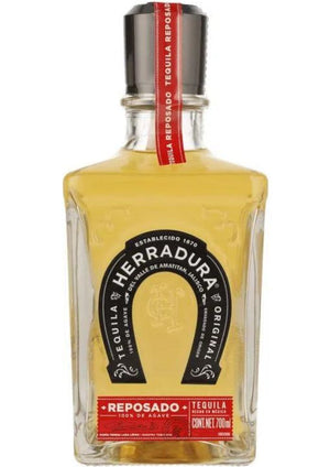 Botella cuadrada con tapón negro, que contiene un líquido ámbar en el interior, así como un sello rojo y etiqueta negra que dice Tequila Herradura. 